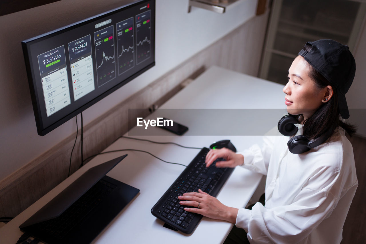 High angle concentrated asian female working on computer with charts showing dynamic of changes in value of cryptocurrency at convenient workplace