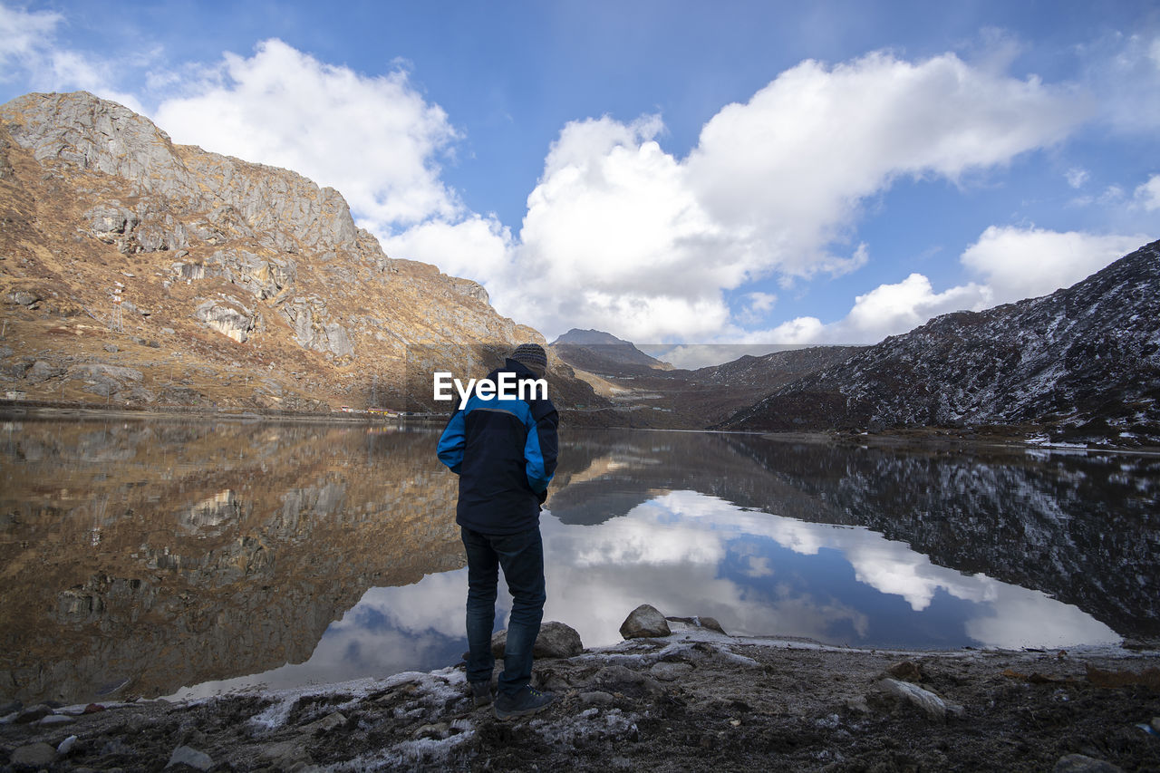 Rear view of mature man standing at lakeshore against cloudy sky