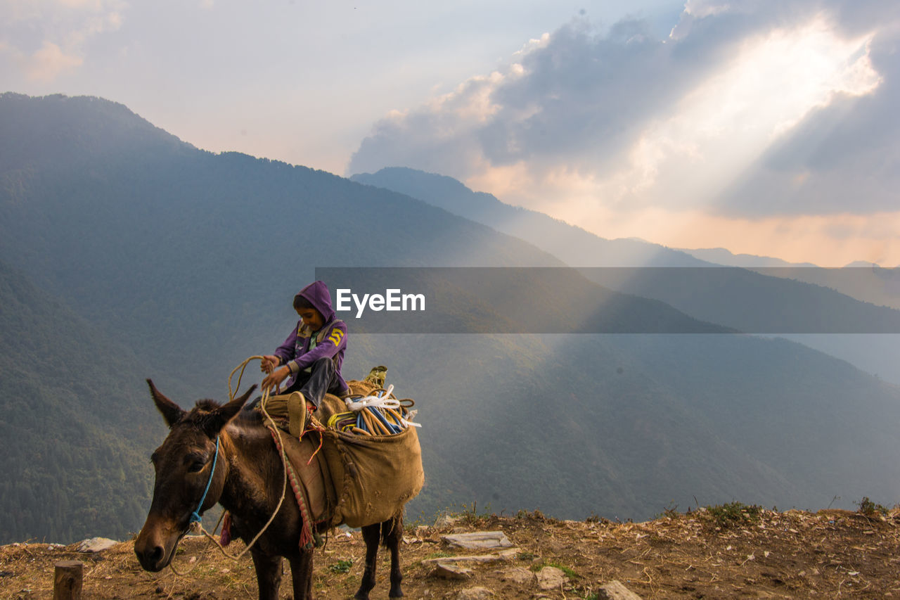 Boy riding horse on mountain against cloudy sky