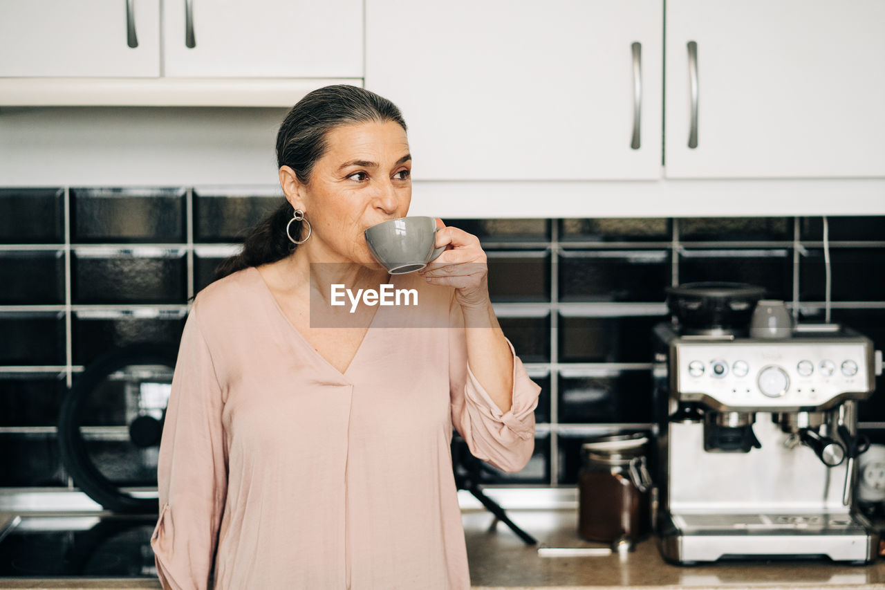 Cheerful middle aged female with mug of hot drink standing at kitchen counter with white cupboards and modern coffee machine at home