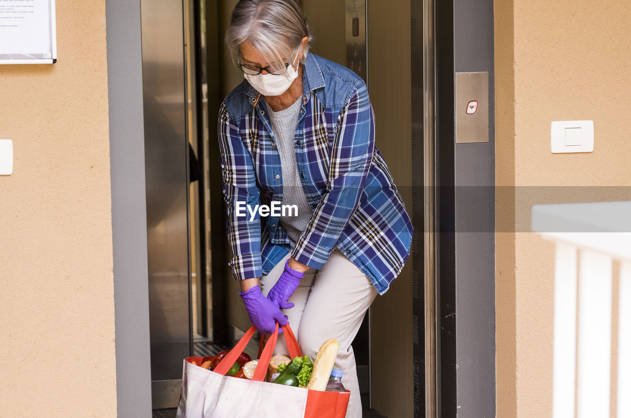 Senior woman wearing mask holding food in bag standing by elevator