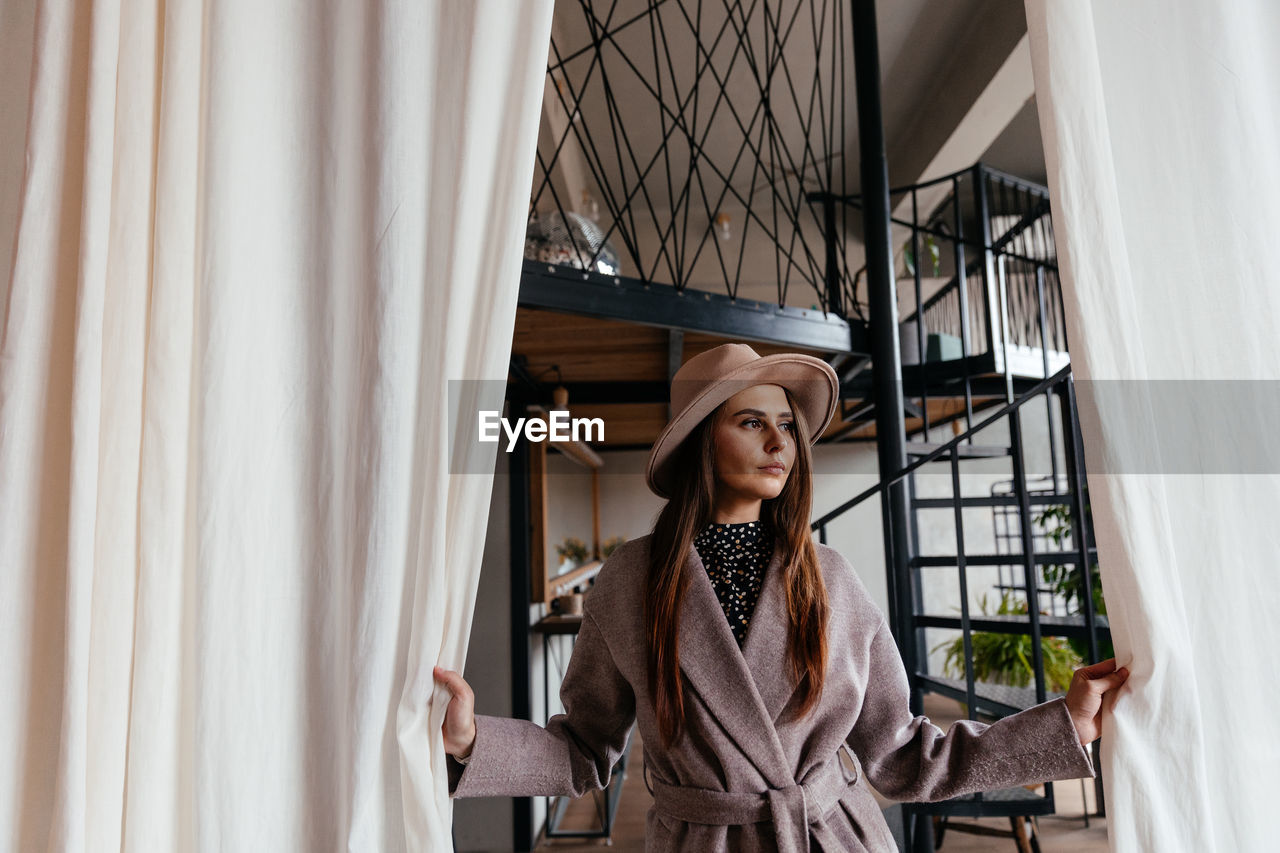 A girl in a beige hat and a beige coat stands indoors and pushes the white curtain with her hands