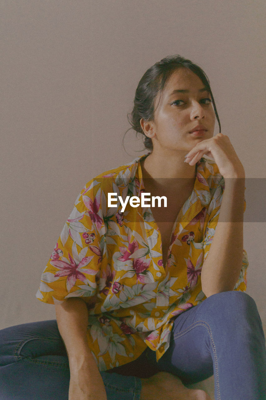 PORTRAIT OF YOUNG WOMAN LOOKING AWAY WHILE SITTING AGAINST WALL AT HOME