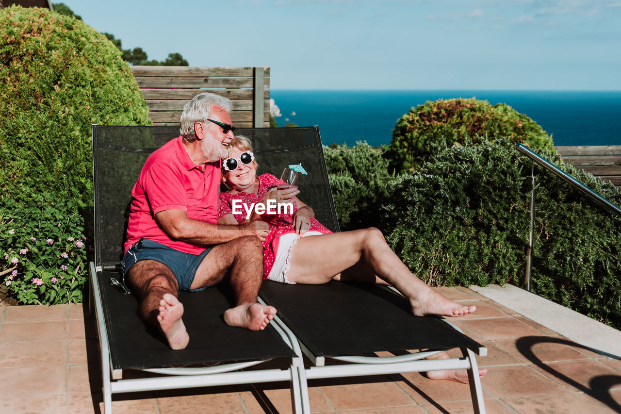 High angle of relaxed senior couple lying on loungers on terrace and drinking refreshing tropical cocktails while enjoying summer holiday