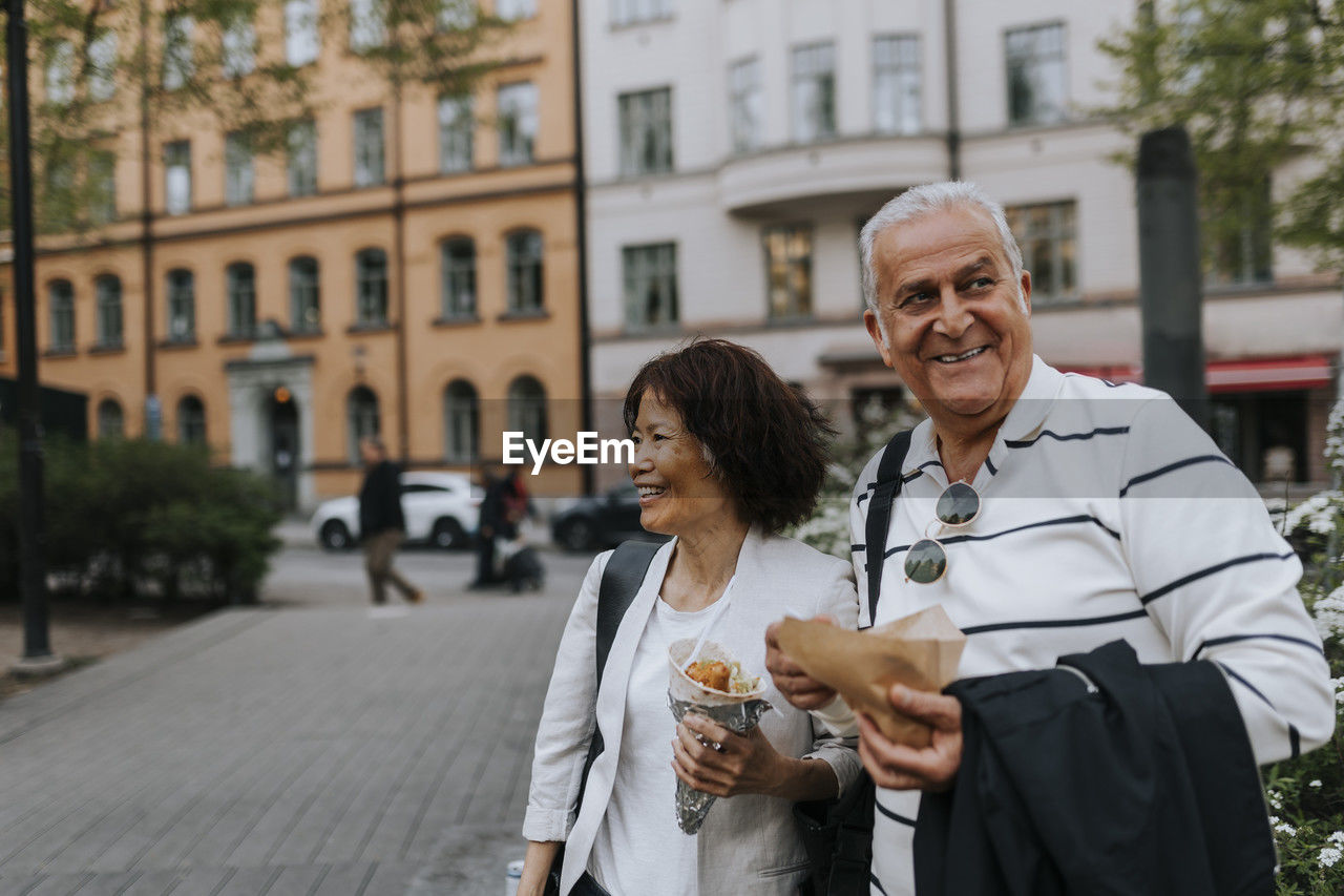 Smiling male and female senior friends looking away while holding snacks at street