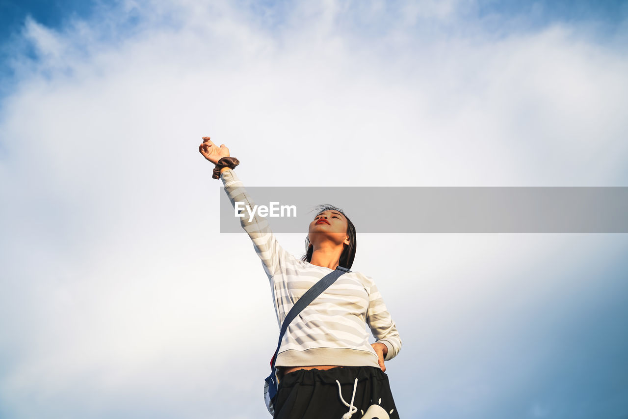 Low angle view of woman with hand raised standing against cloudy sky