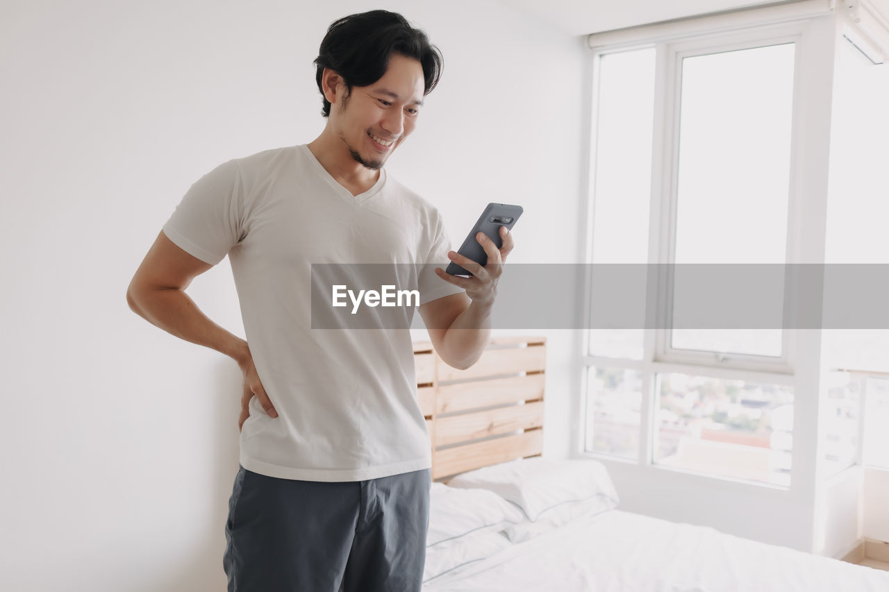Young man using mobile phone while standing on bed