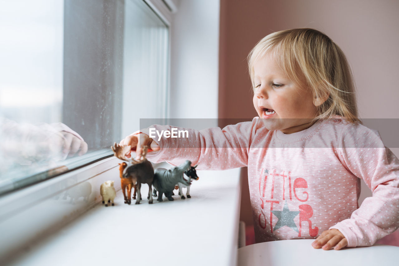 Little girl toddler playing with animal toys on window sill in children's room at home