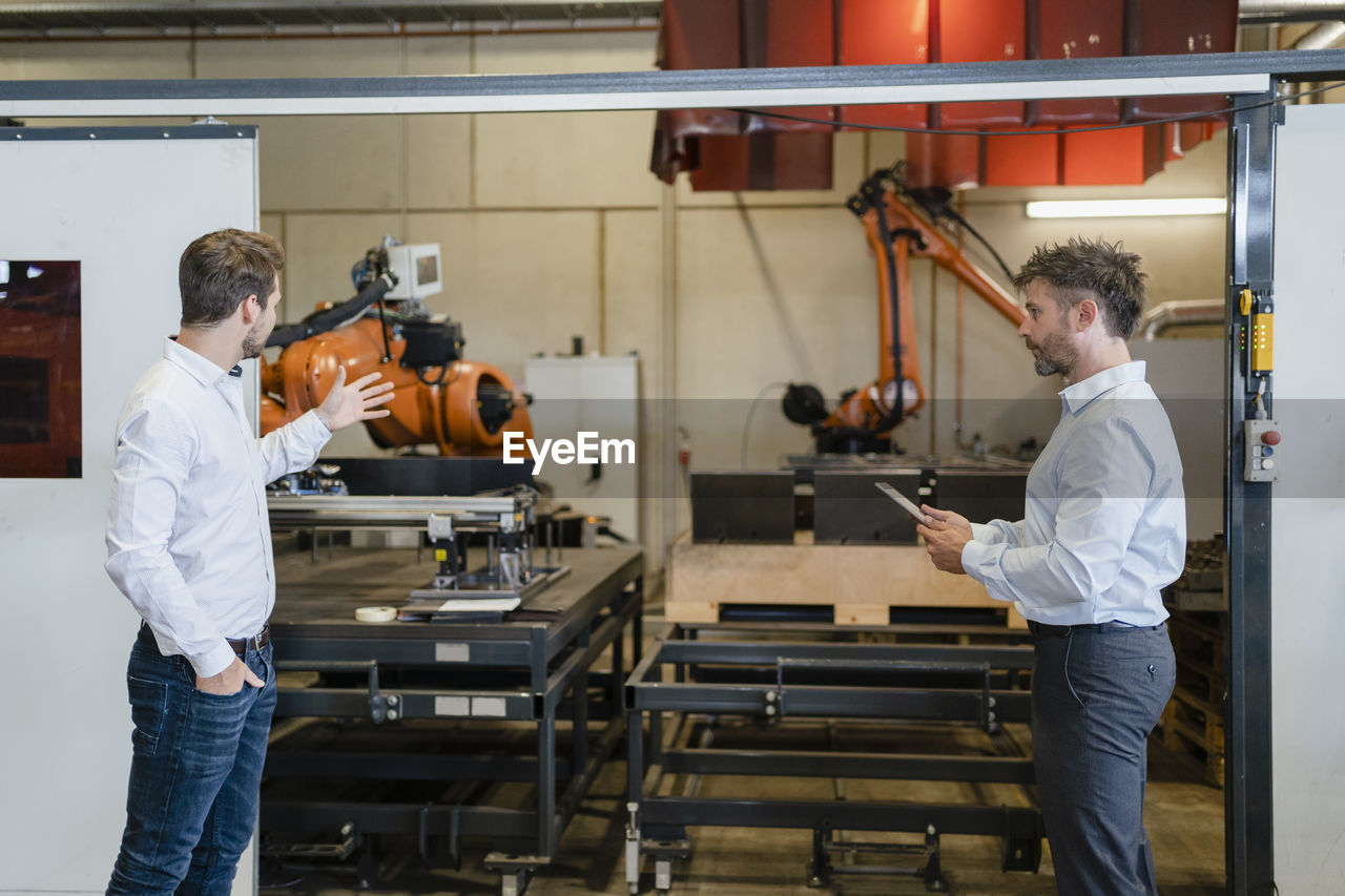 Businessman having discussion with colleague while standing by robotic arm machine at factory