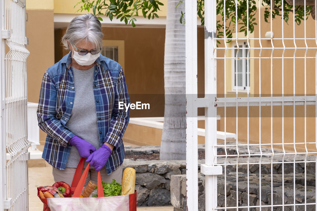 Senior woman wearing mask holding food standing by gate