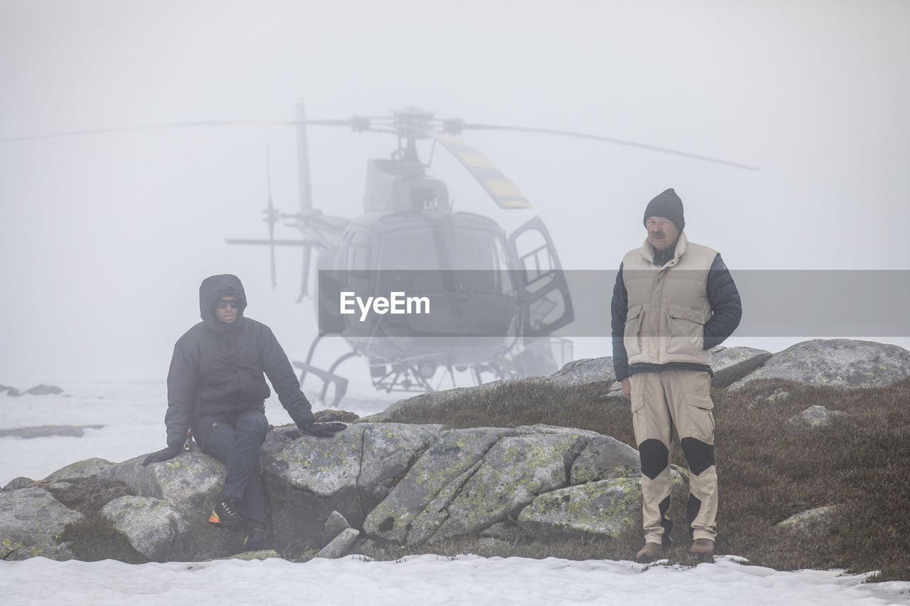 Helicopter pilot and passenger wait on mountain for weather to clear.