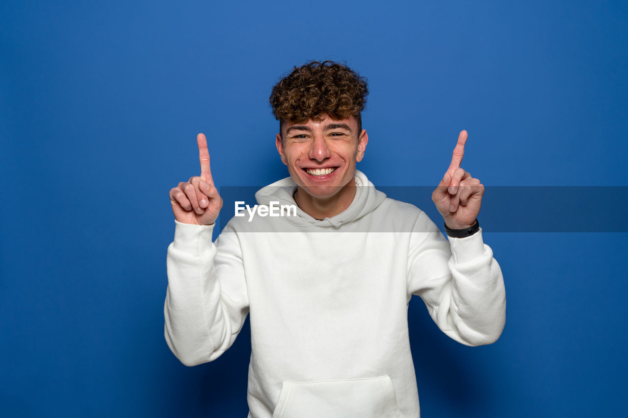 sign language, gesturing, finger, one person, blue, thumbs up, studio shot, hand sign, hand, smiling, person, colored background, happiness, emotion, waist up, portrait, men, front view, adult, looking at camera, arm, standing, blue background, clothing, white, indoors, success, cheerful, positive emotion, showing, young adult, communication, casual clothing, pointing, human face, limb, human limb, hand raised, fun, arms raised, smile, teeth, peace sign