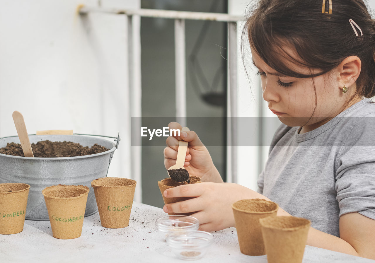 A caucasian girl enthusiastically fills the soil with a wooden spoon into a cardboard glass.