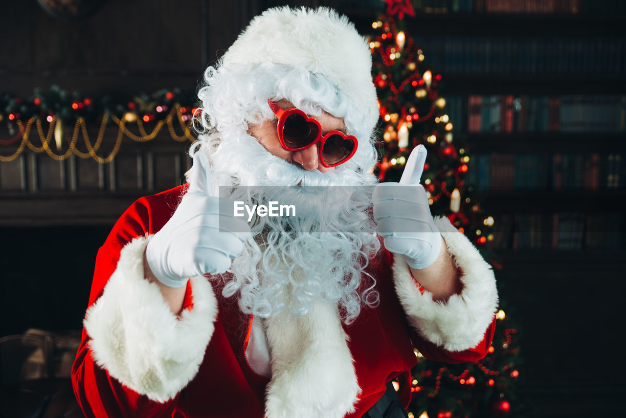 Portrait of santa clause wearing sunglasses standing against illuminated christmas tree