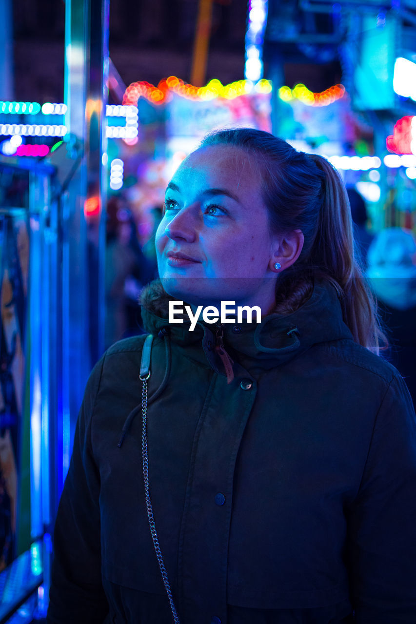 Close-up of young woman standing against illuminated amusement ride at night