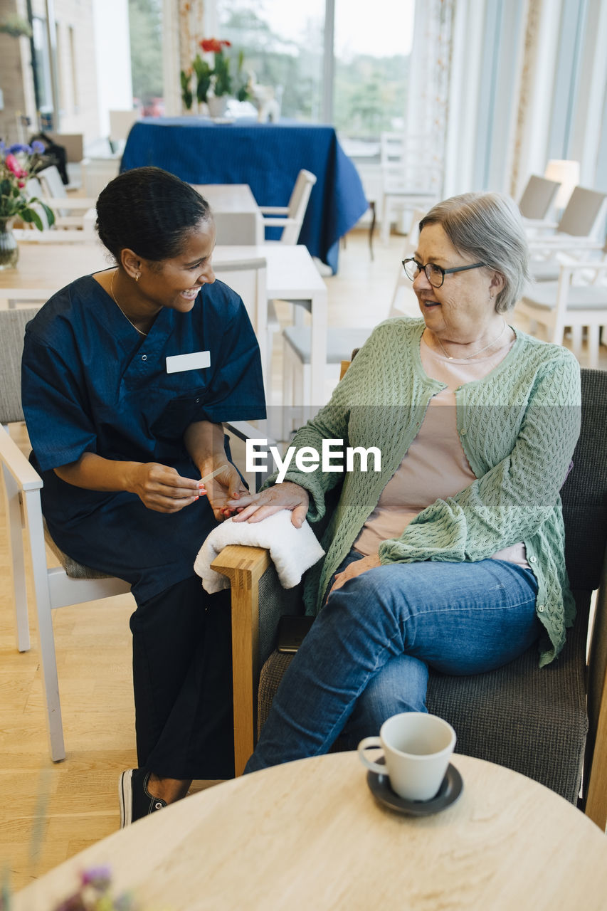 Senior woman looking at young female caretaker filing her nails in retirement home