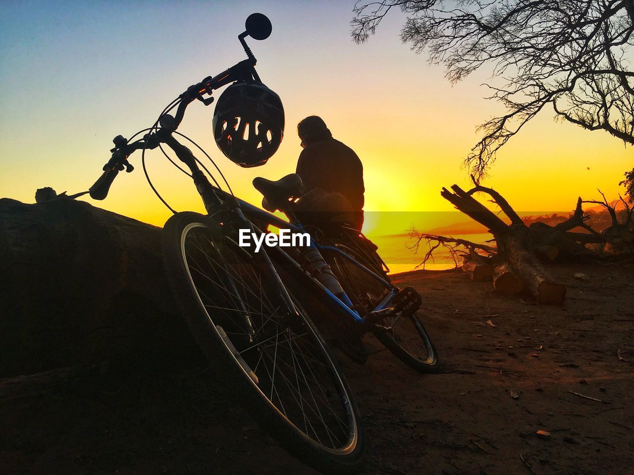 Silhouette man sitting by bicycle against orange sky during sunset