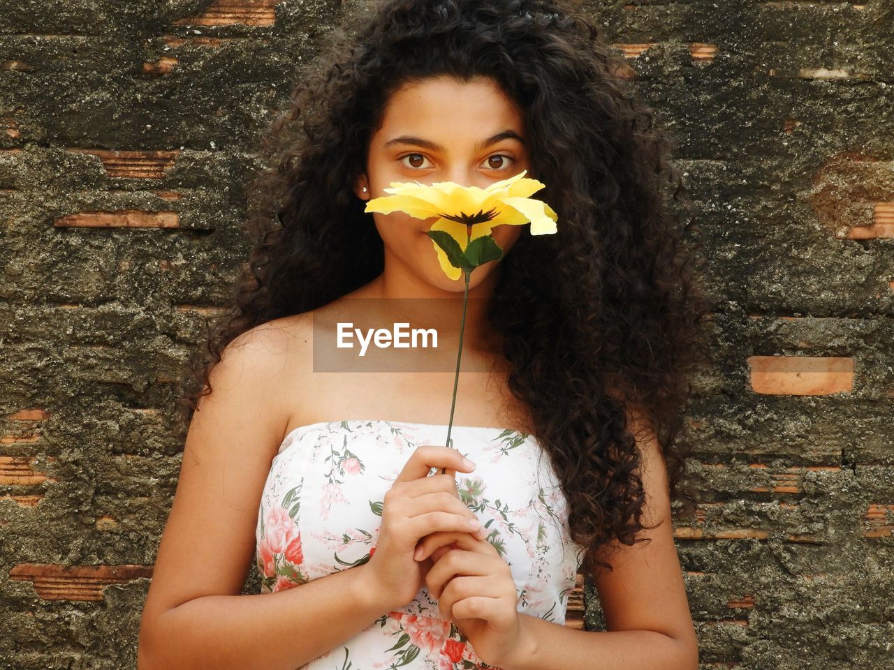 Portrait of beautiful teenage girl with artificial flower standing against brick wall