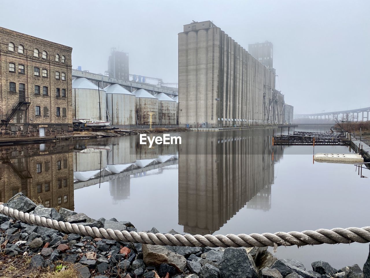 Industrial buildings in a shipyard reflected in water on an overcast day. 