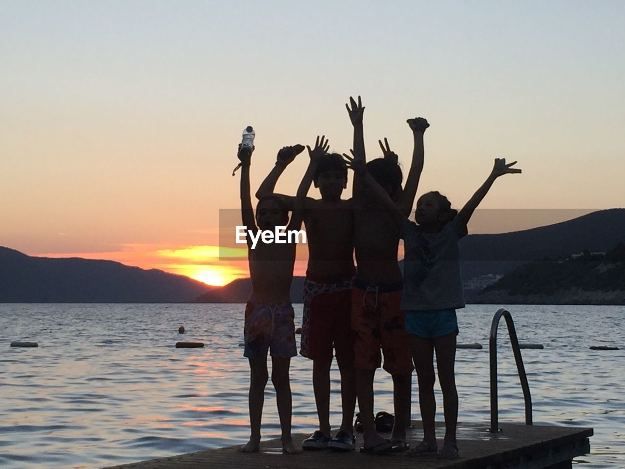 Cheerful friends with arms raised standing on pier at lake during sunset