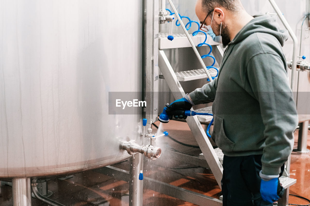 Side view of crop male worker in mask washing valve of metal tank at work in brewing company during pandemic