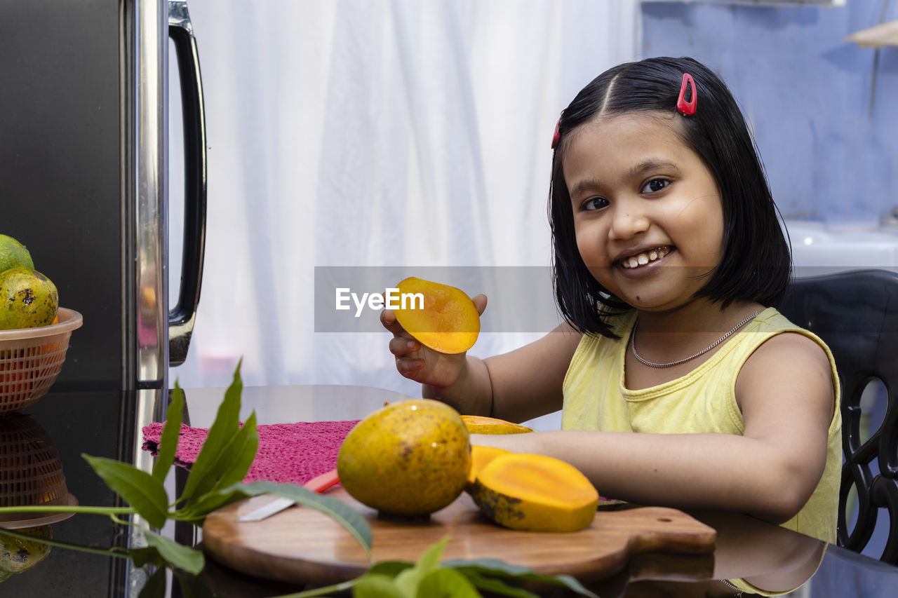 An indian girl child eating mango and smiling at the camera sitting beside a table 