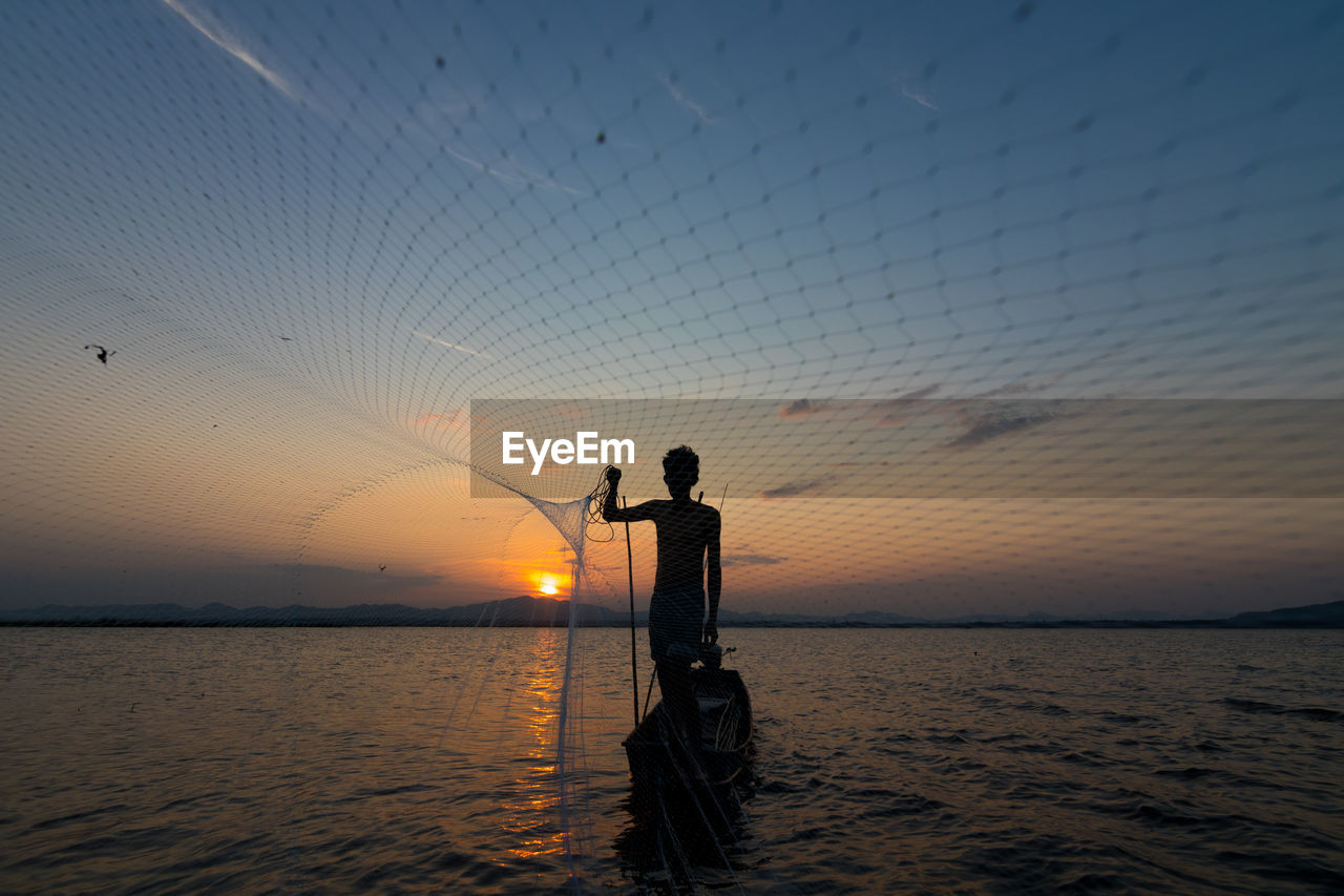 Silhouette of man throwing fishing net while standing in boat on sea against sky during sunset