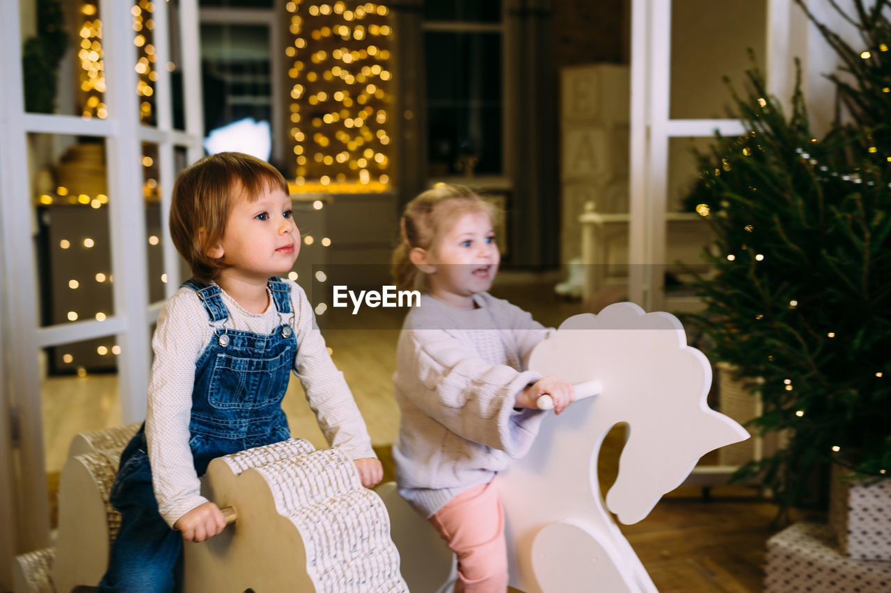 Little children ride toy horses in decorated christmas interior.