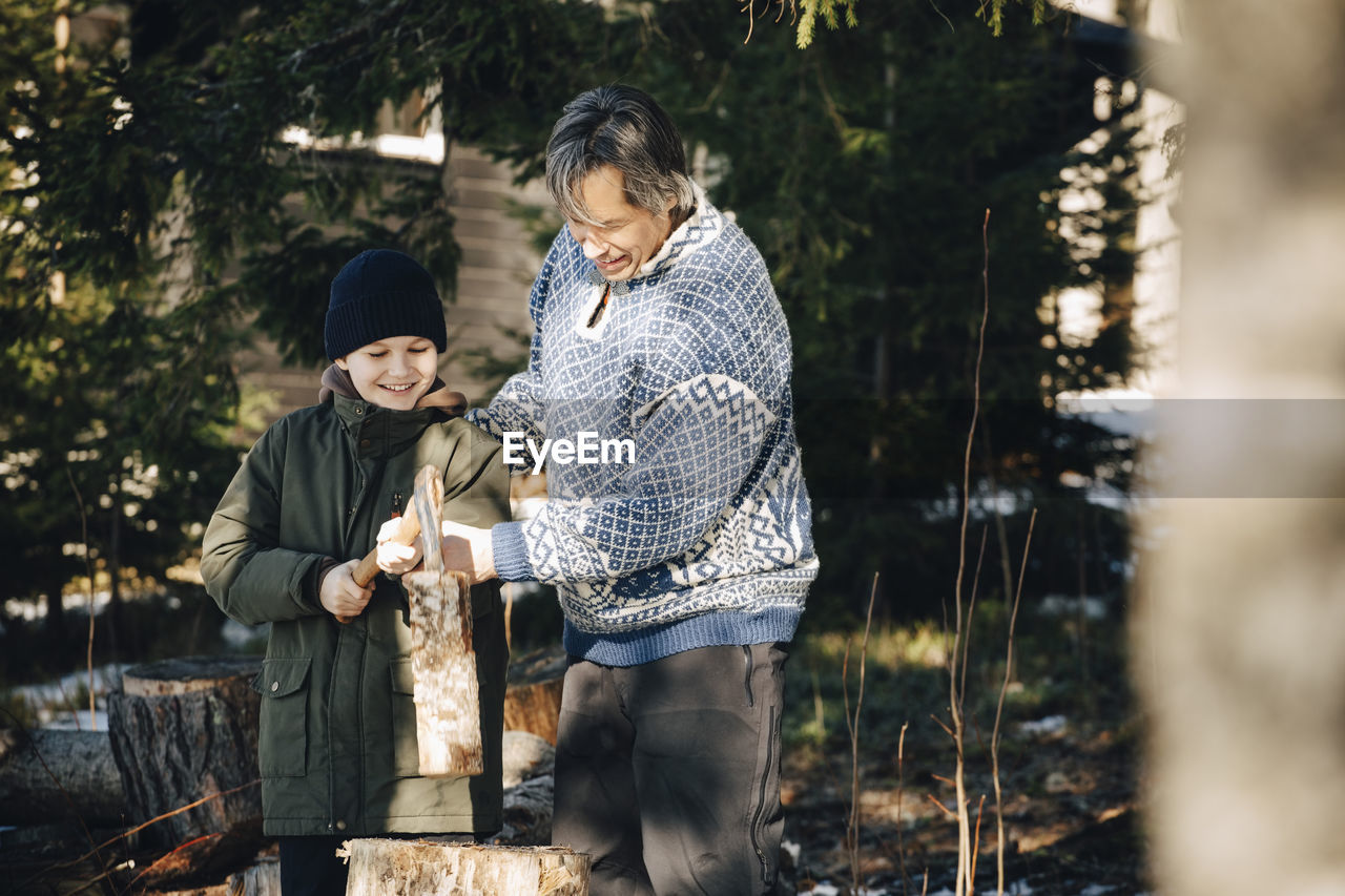 Smiling mature man teaching son to cut log with axe in forest