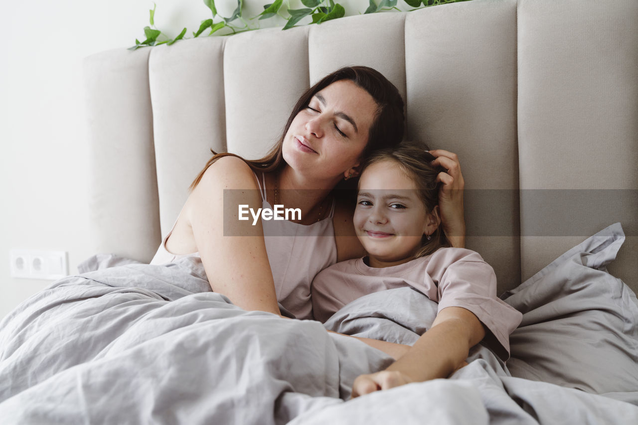 Woman with eyes closed embracing daughter lying on bed at home