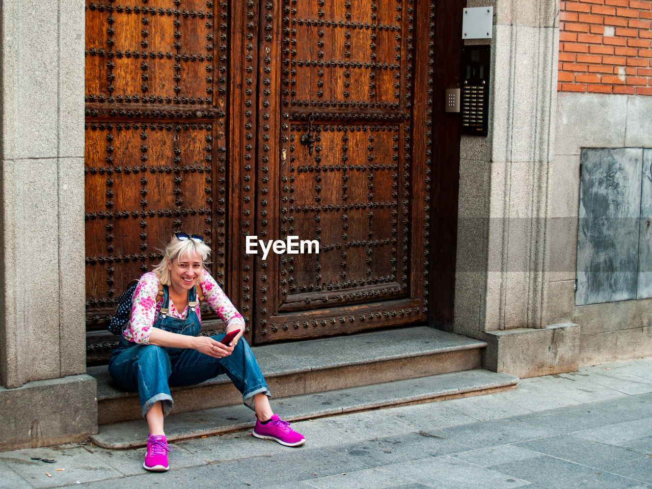 Full length of smiling woman holding mobile phone while sitting at entrance