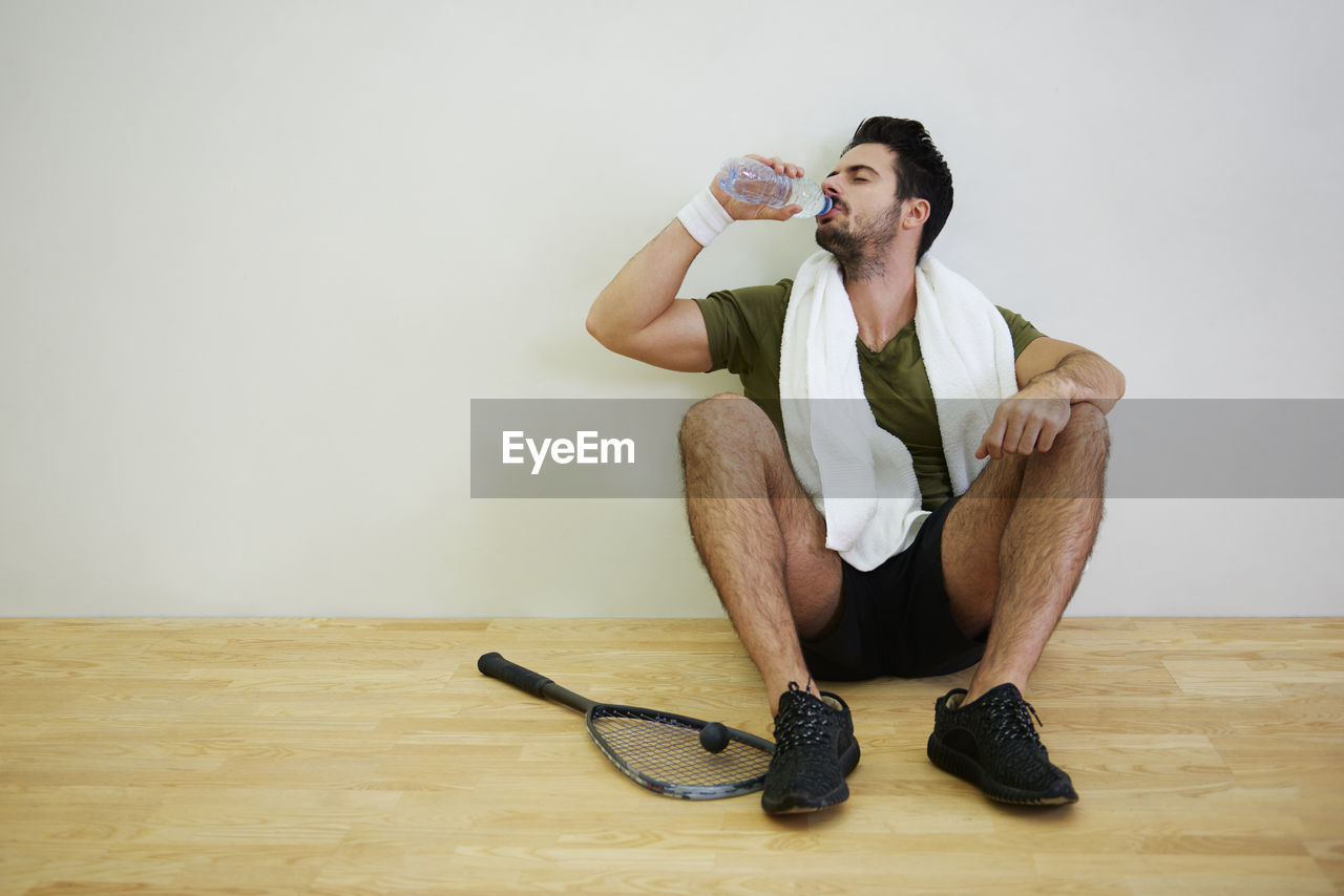 Full length of man drinking water while sitting on floor against wall