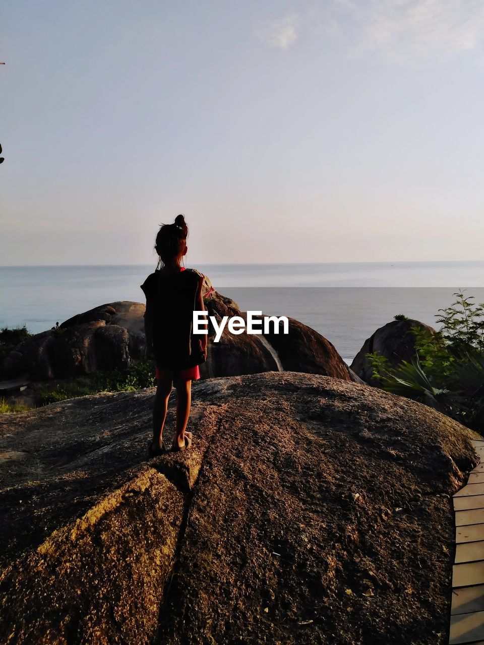 MAN STANDING ON ROCK LOOKING AT SEA SHORE AGAINST SKY