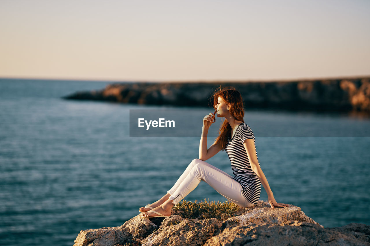 MAN SITTING ON ROCK AT SEA SHORE AGAINST SKY