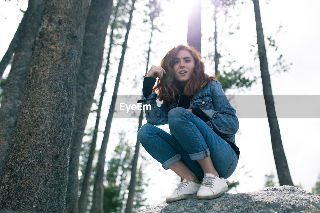 Low angle portrait of smiling young woman crouching on rock in forest