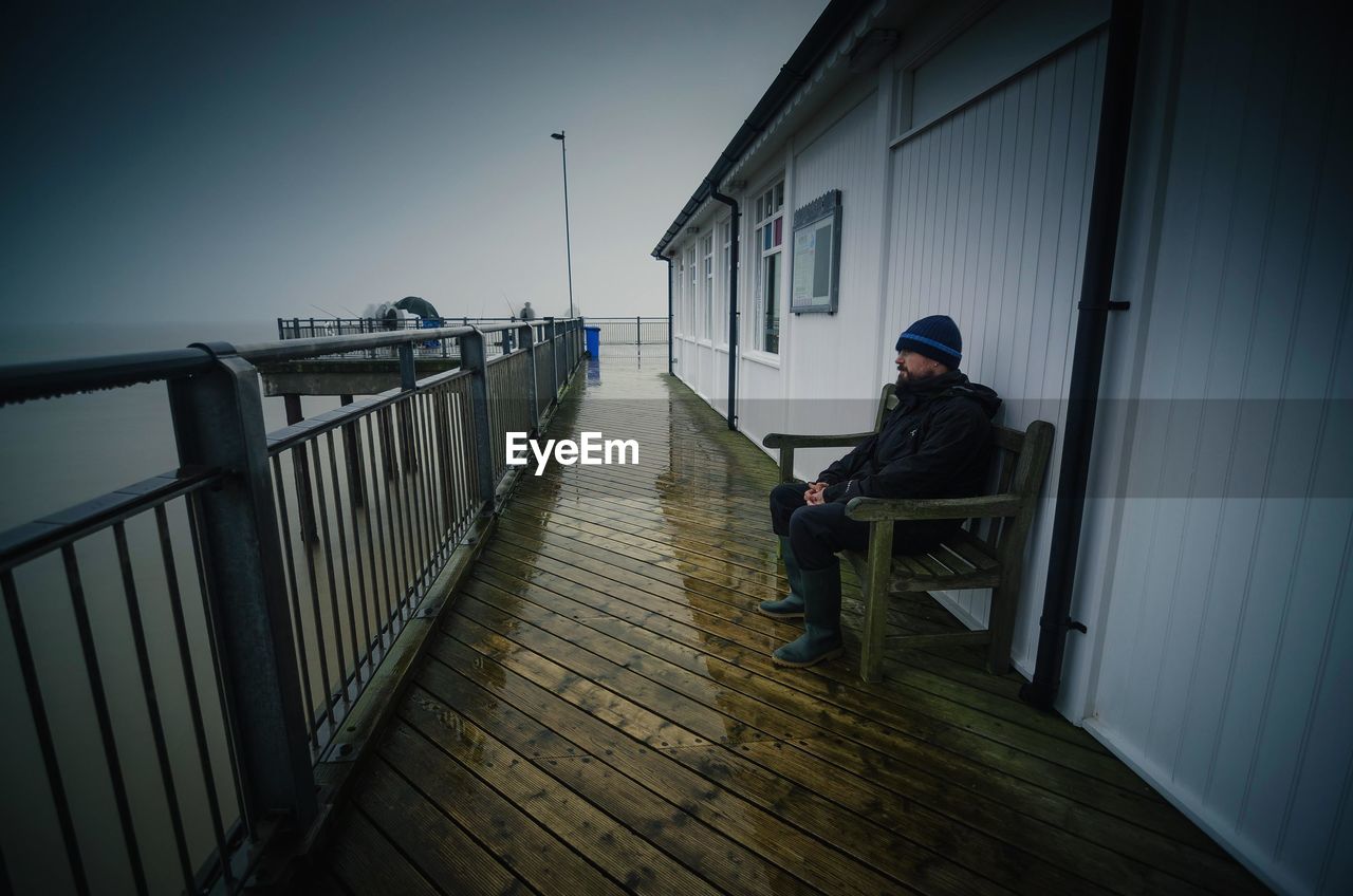 Man sitting on bench by wet walkway and railing at pier