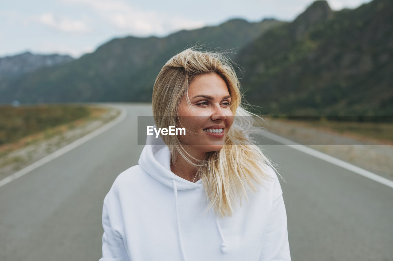 Beautiful smiling blonde young woman traveler in white hoodie on road, trip to mountains, altai