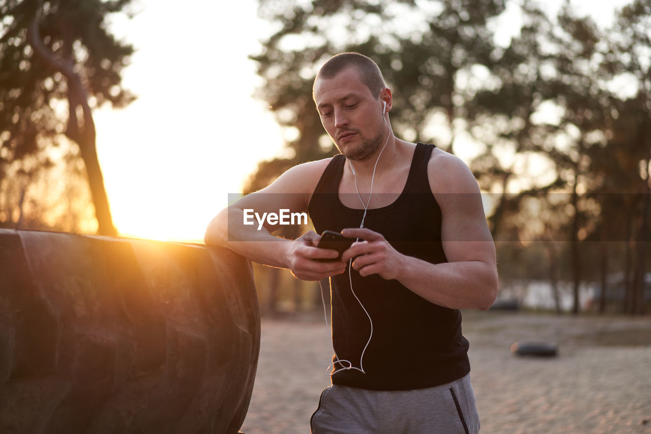 Young man using mobile phone while standing by tire at sunset