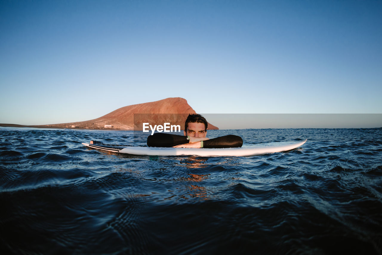 Happy male resting surfer on surfboard in wavy sea looking at camera against mount in daytime