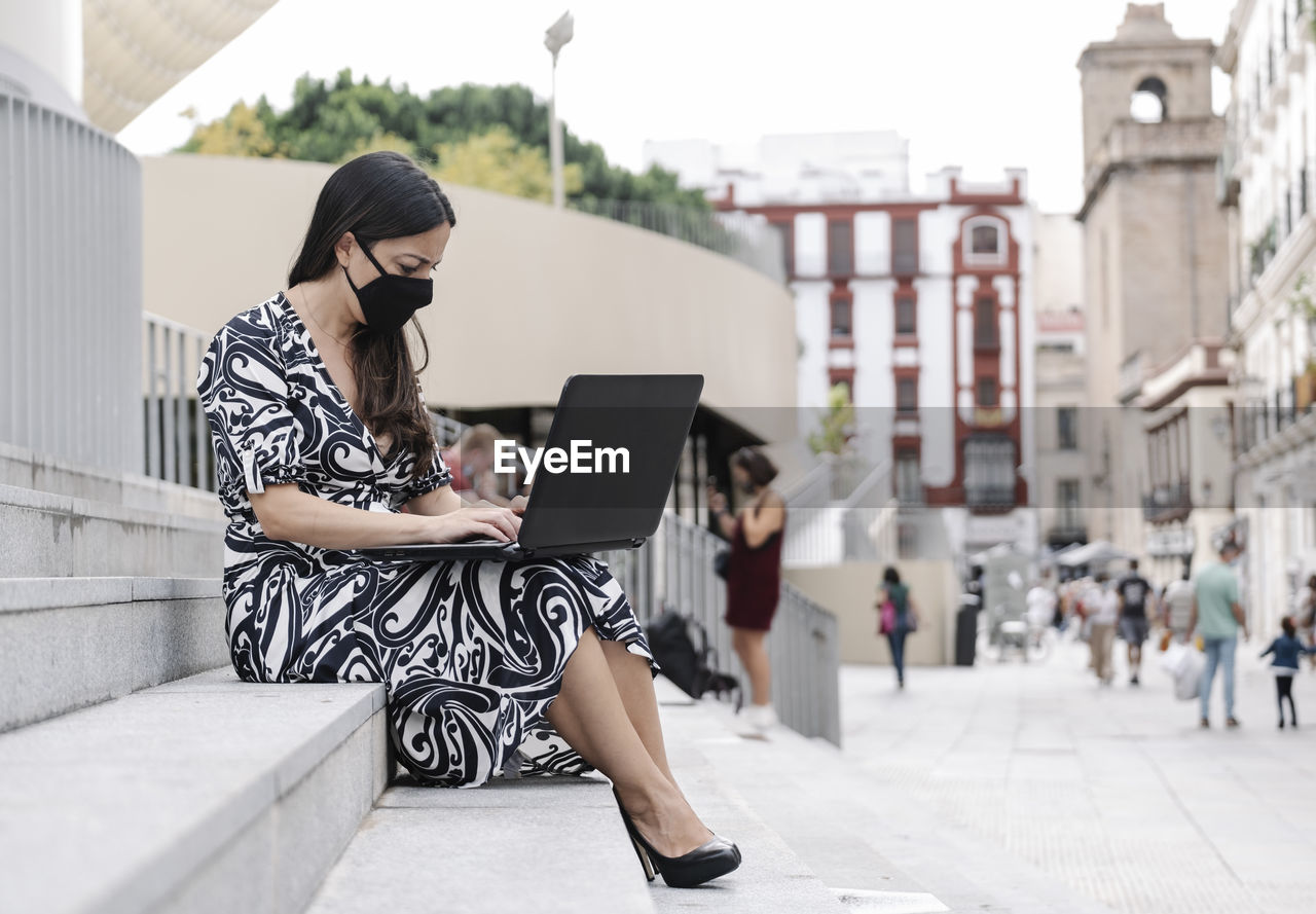 Full length of businesswoman wearing mask using laptop while sitting on steps outdoors