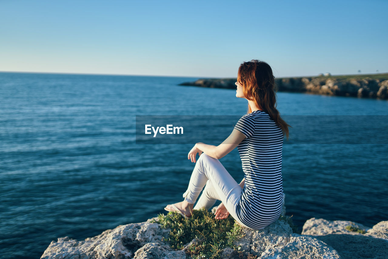 Woman sitting on rock looking at sea against sky