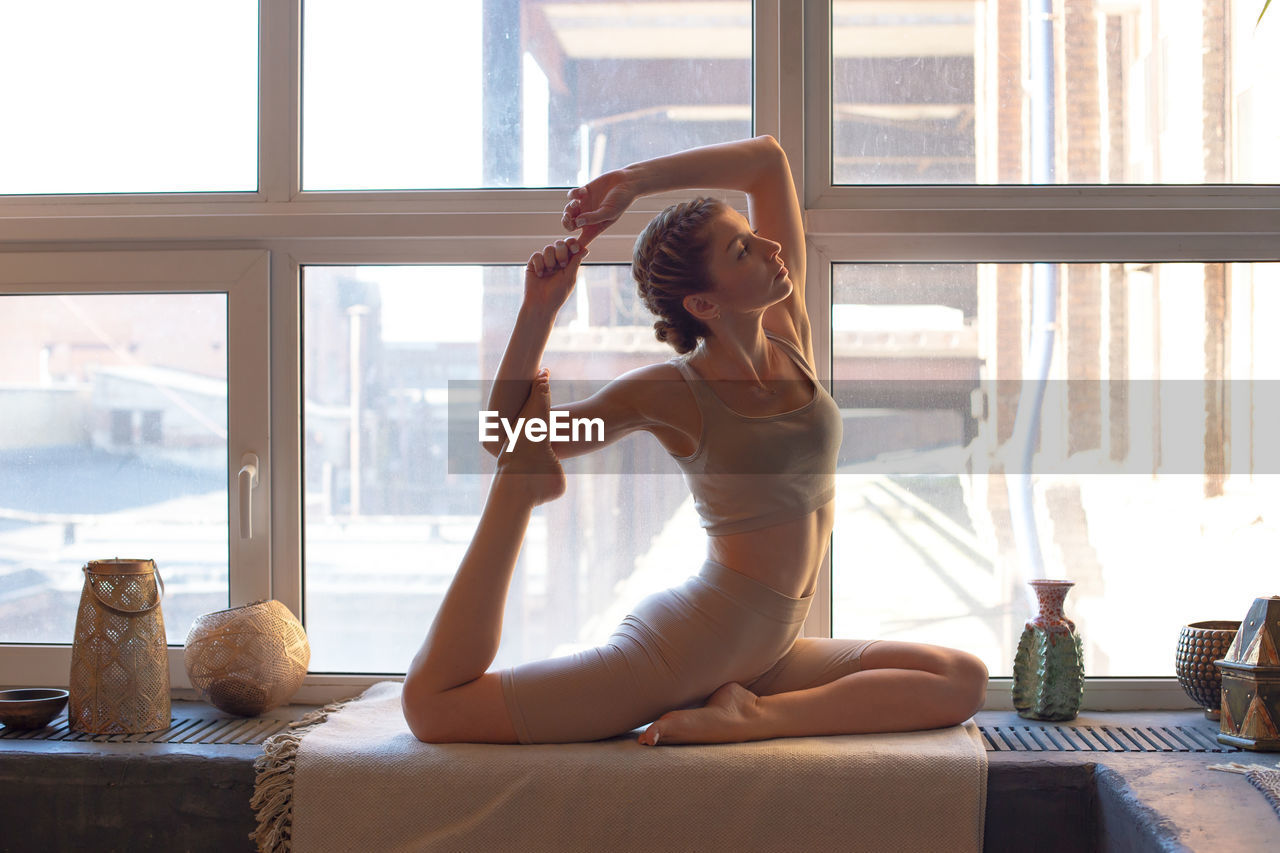 A slender and beautiful woman in a beige top and leggings sits by a large window in a yoga pose