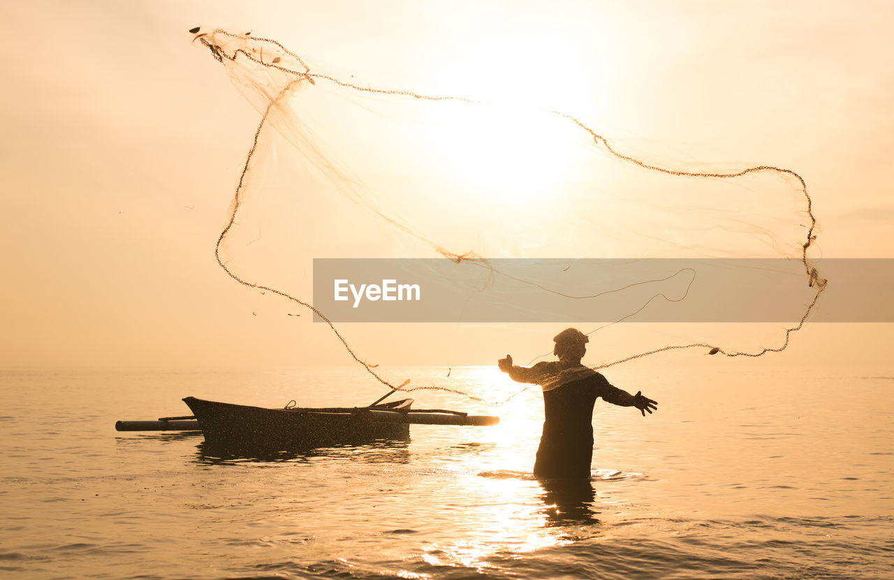 Silhouette fisherman fishing in sea against orange sky