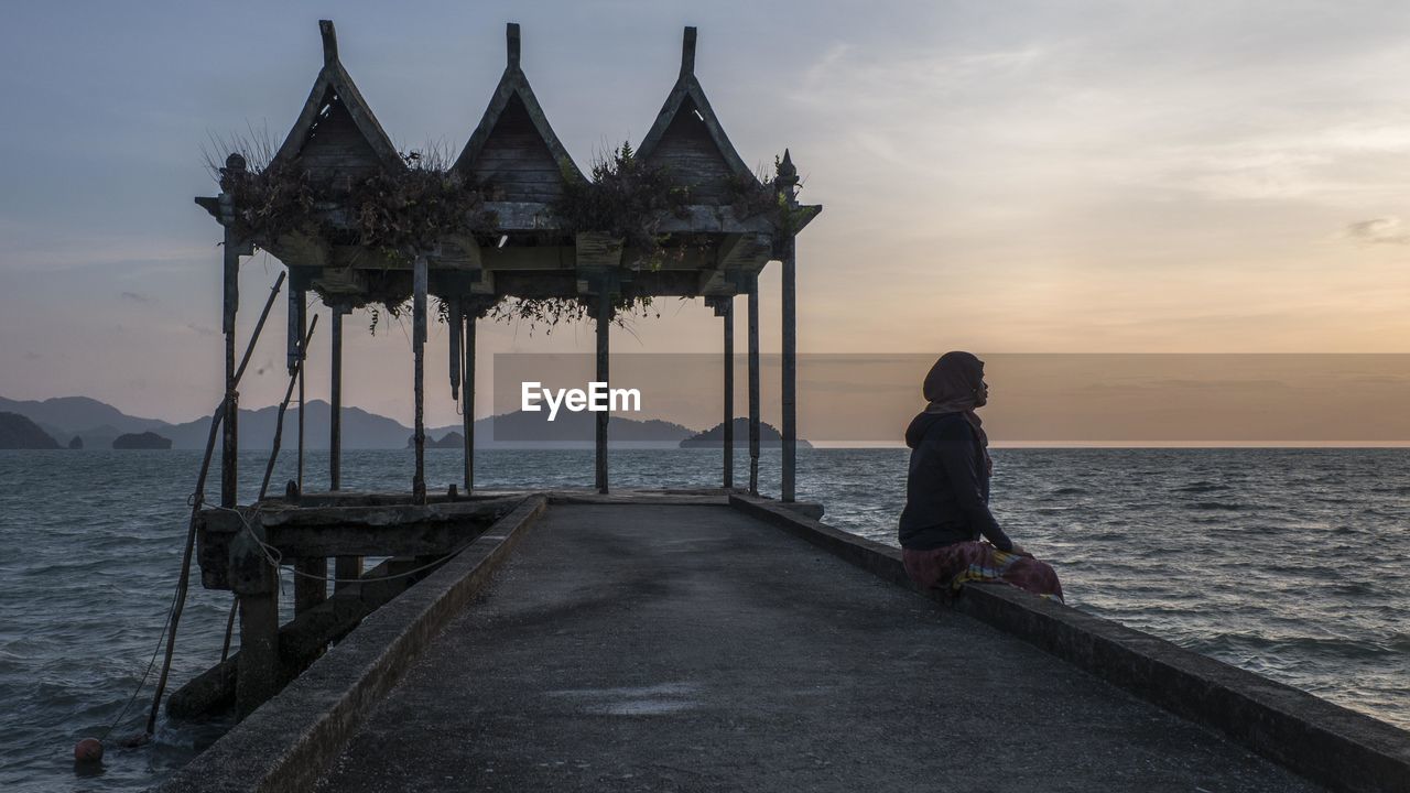 REAR VIEW OF MAN STANDING ON PIER AT SEA AGAINST SKY