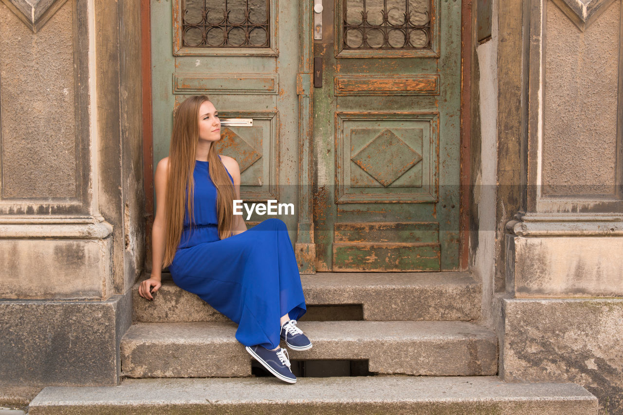 Full length of thoughtful beautiful woman sitting on steps by closed weathered doors