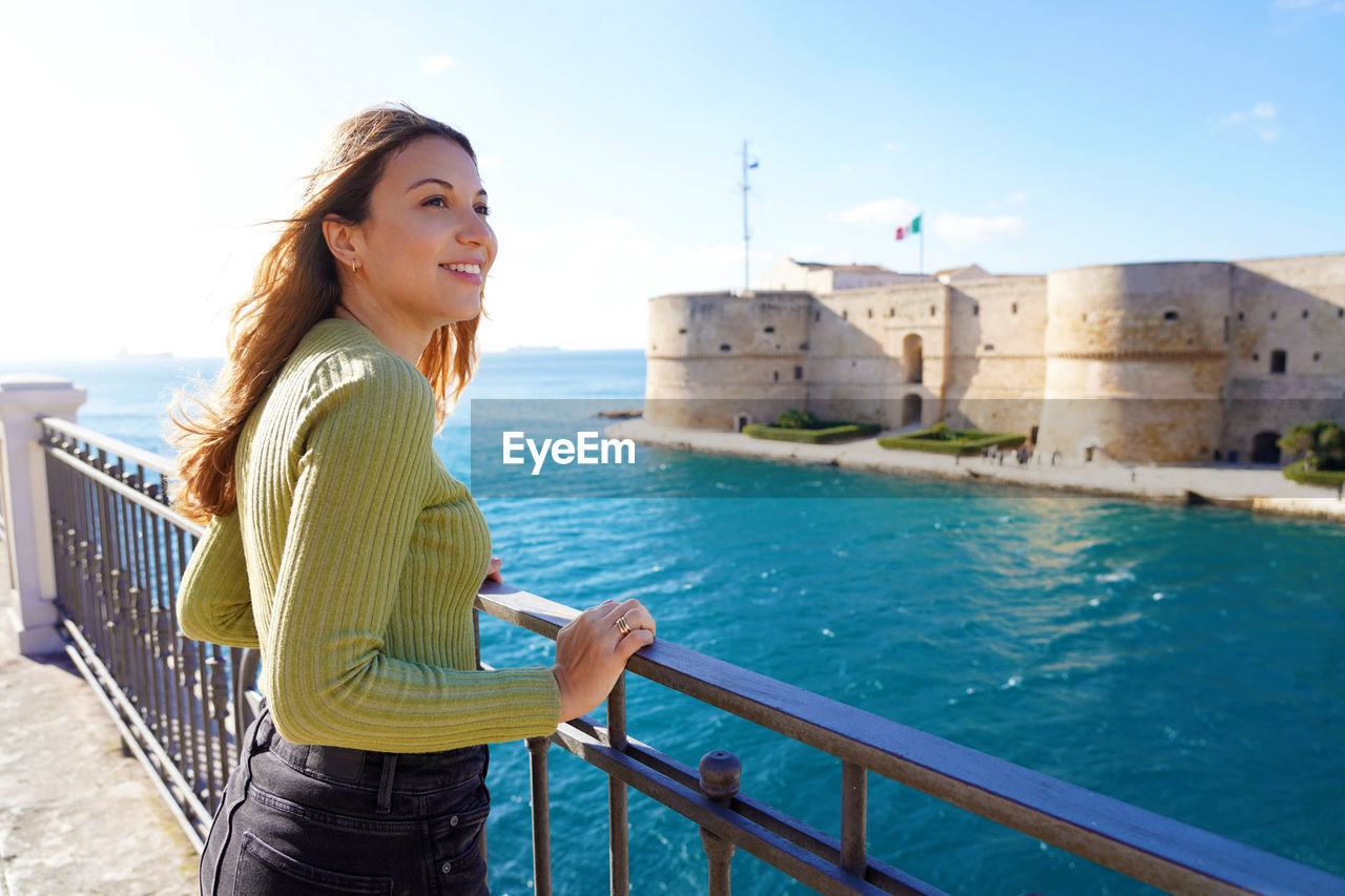 Portrait of smiling relaxed traveler woman looking taranto city from seafront, apulia, italy