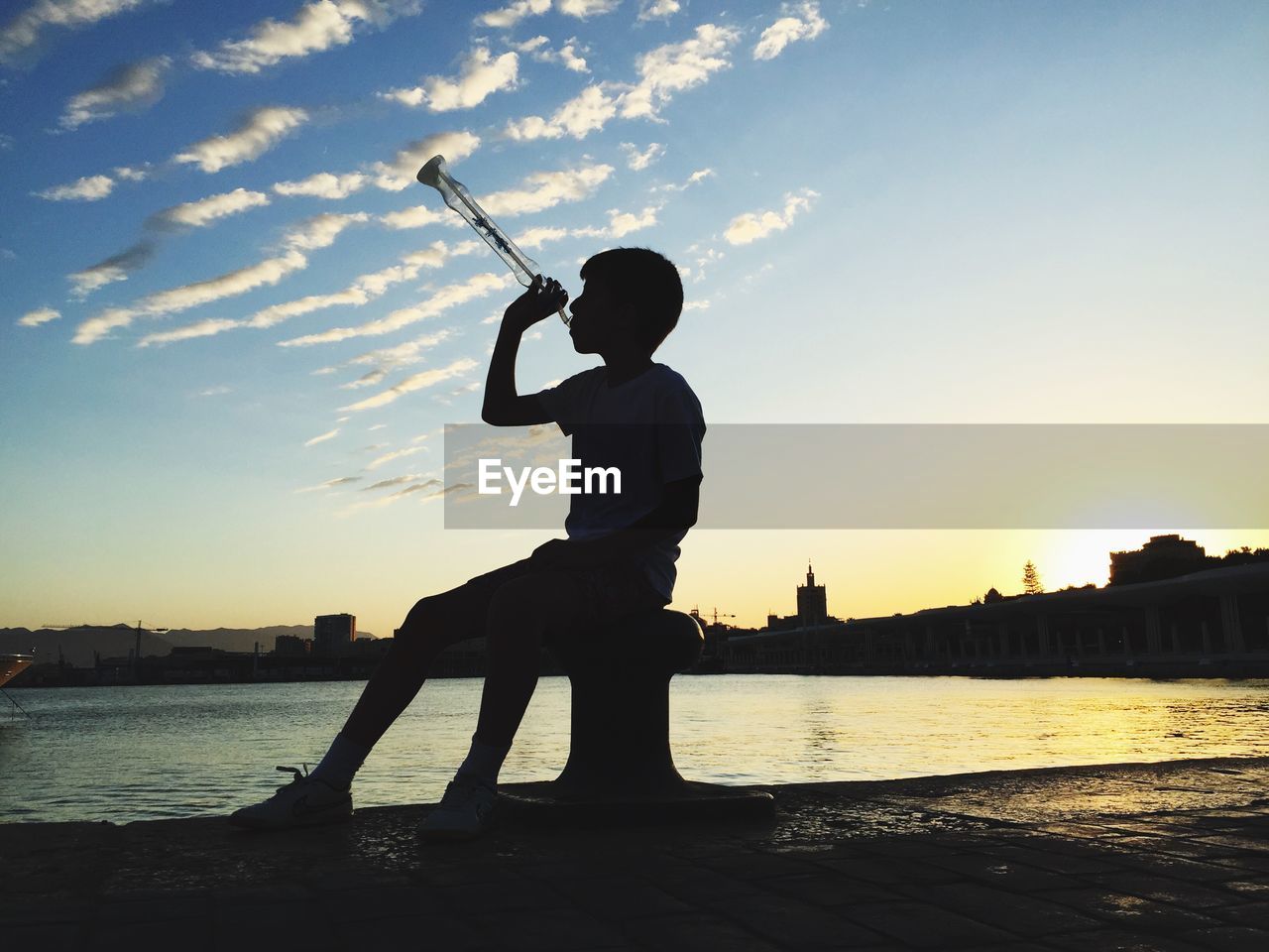 Boy with drink while sitting on bollard at pier during sunset