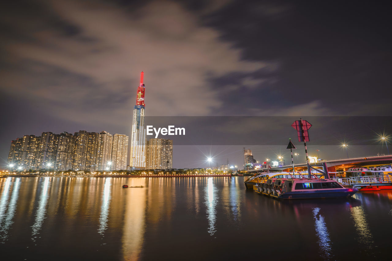 ILLUMINATED BUILDINGS BY RIVER AGAINST SKY IN CITY