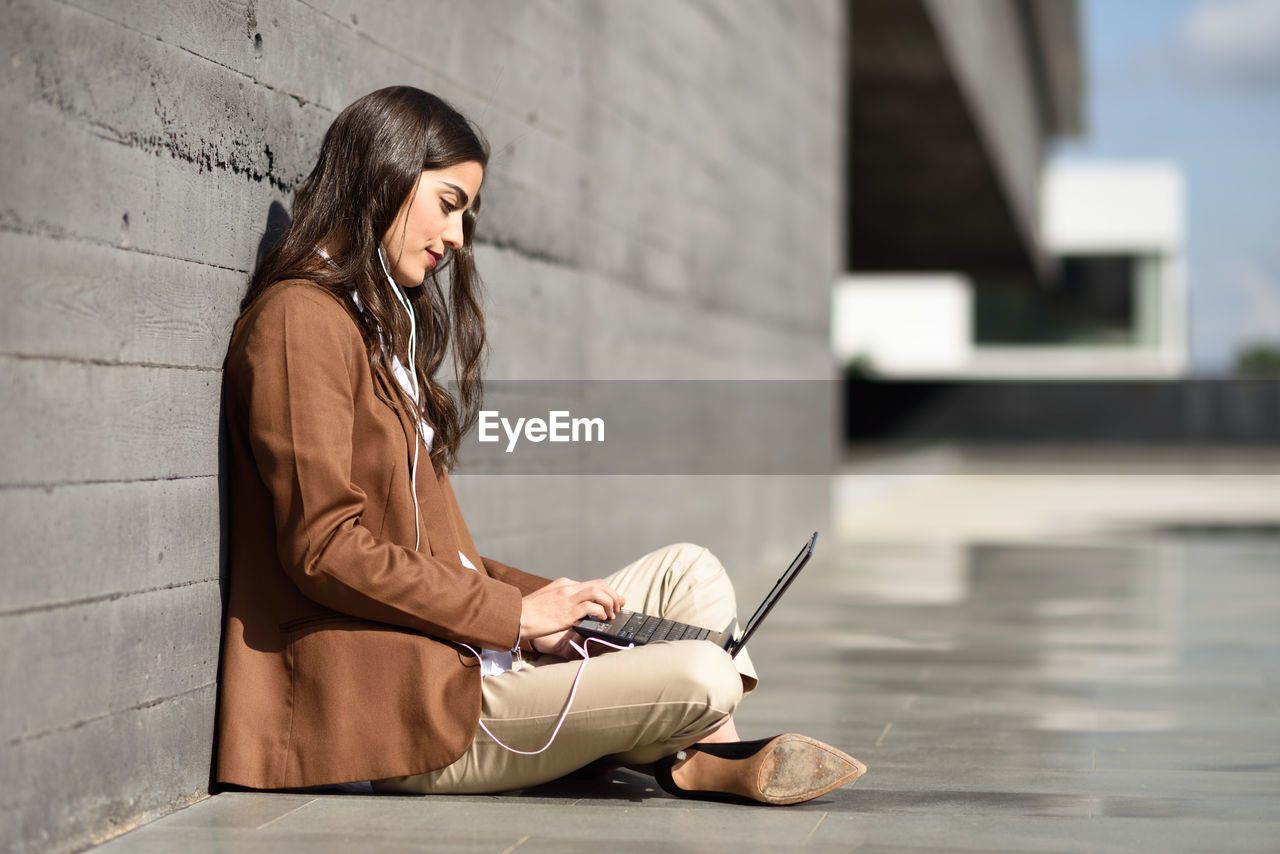 Young businesswoman using laptop while sitting against wall