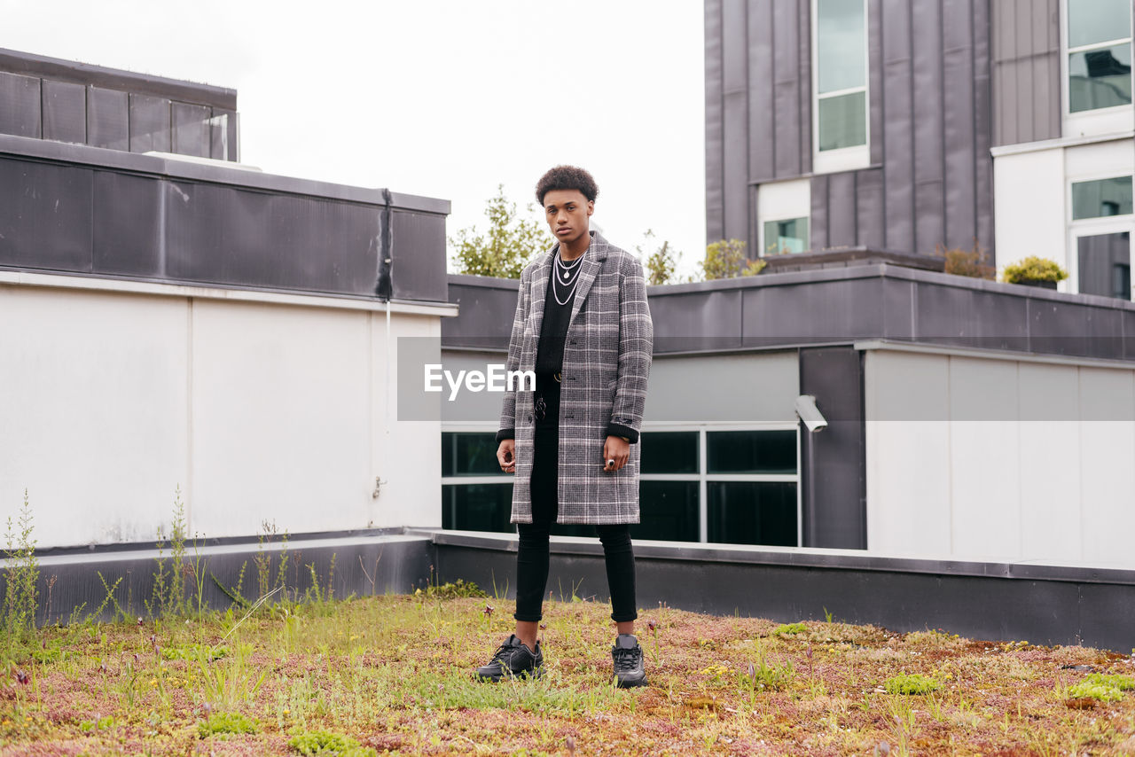 Serious young african american fashionable guy in trendy checkered coat and tight pants looking at camera while standing against modern buildings in city